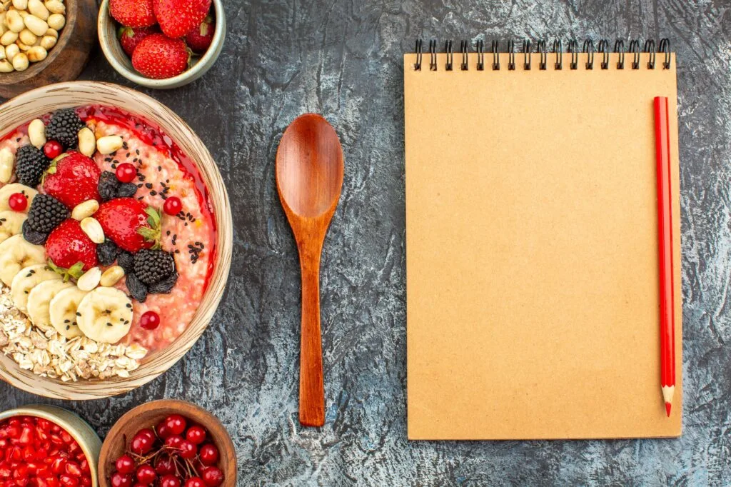 A bowl of oatmeal topped with strawberries blackberries banana slices red currants nuts and seeds is placed next to a wooden spoon and a blank notepad with a red pencil on a textured surface